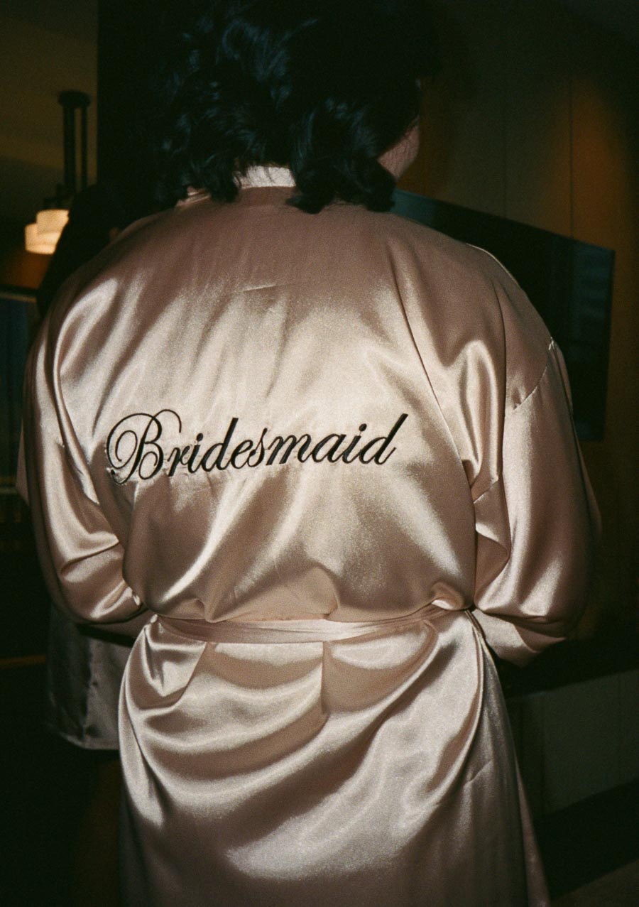 Back of a bridesmaid wearing a satin robe with Bridesmaid embroidered on it, preparing for a wedding ceremony.