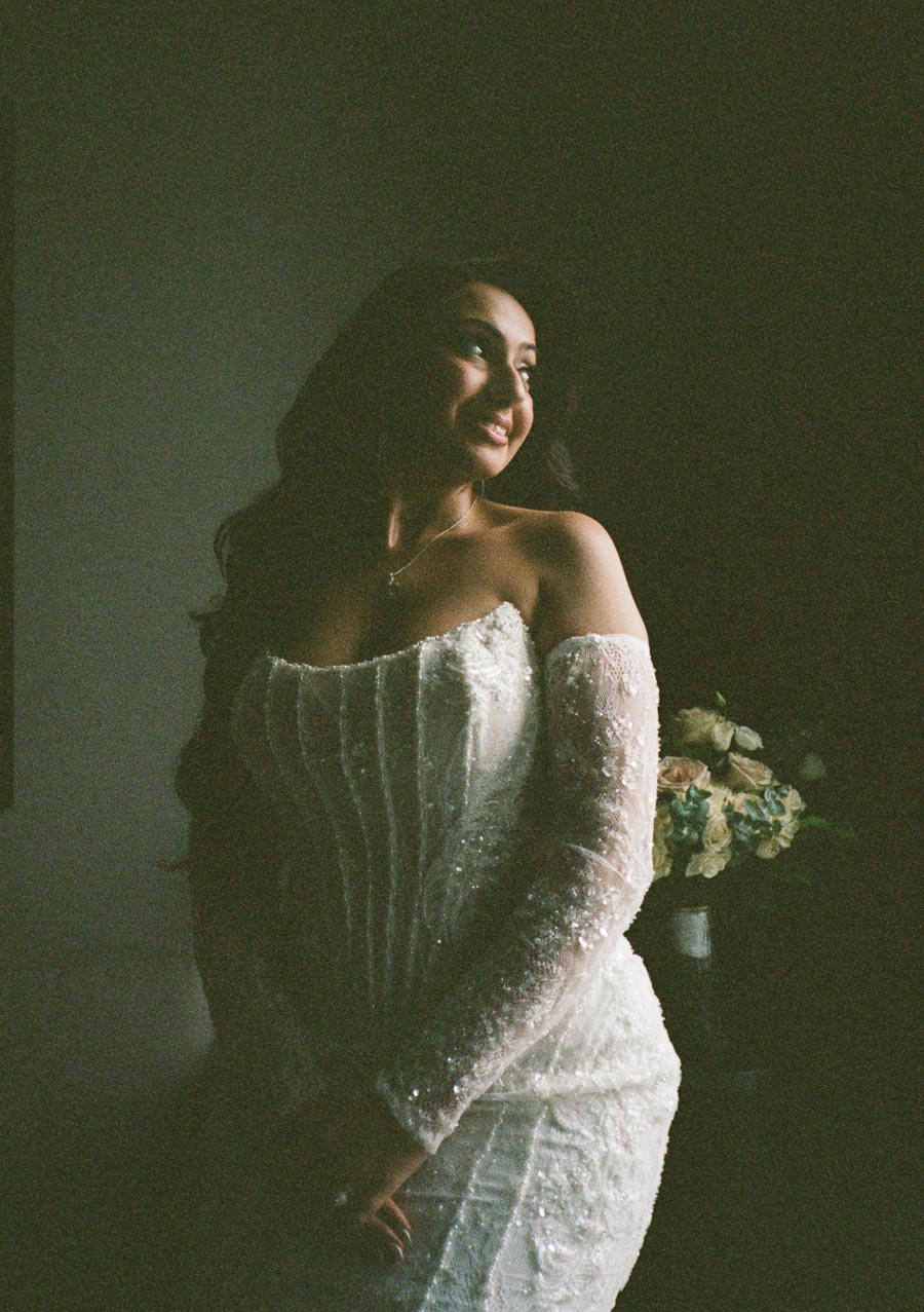 Elegant bride in an off-shoulder lace wedding gown smiles softly while standing in a softly lit room, bouquet of flowers in the background.