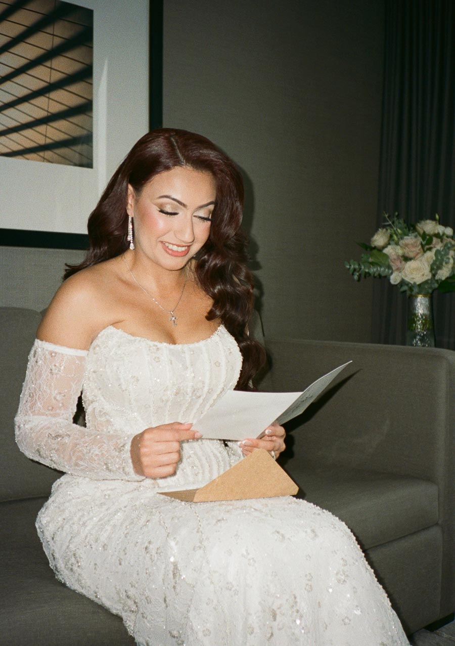 A woman in an elegant white lace gown sits on a sofa, reading a letter with a joyful expression.