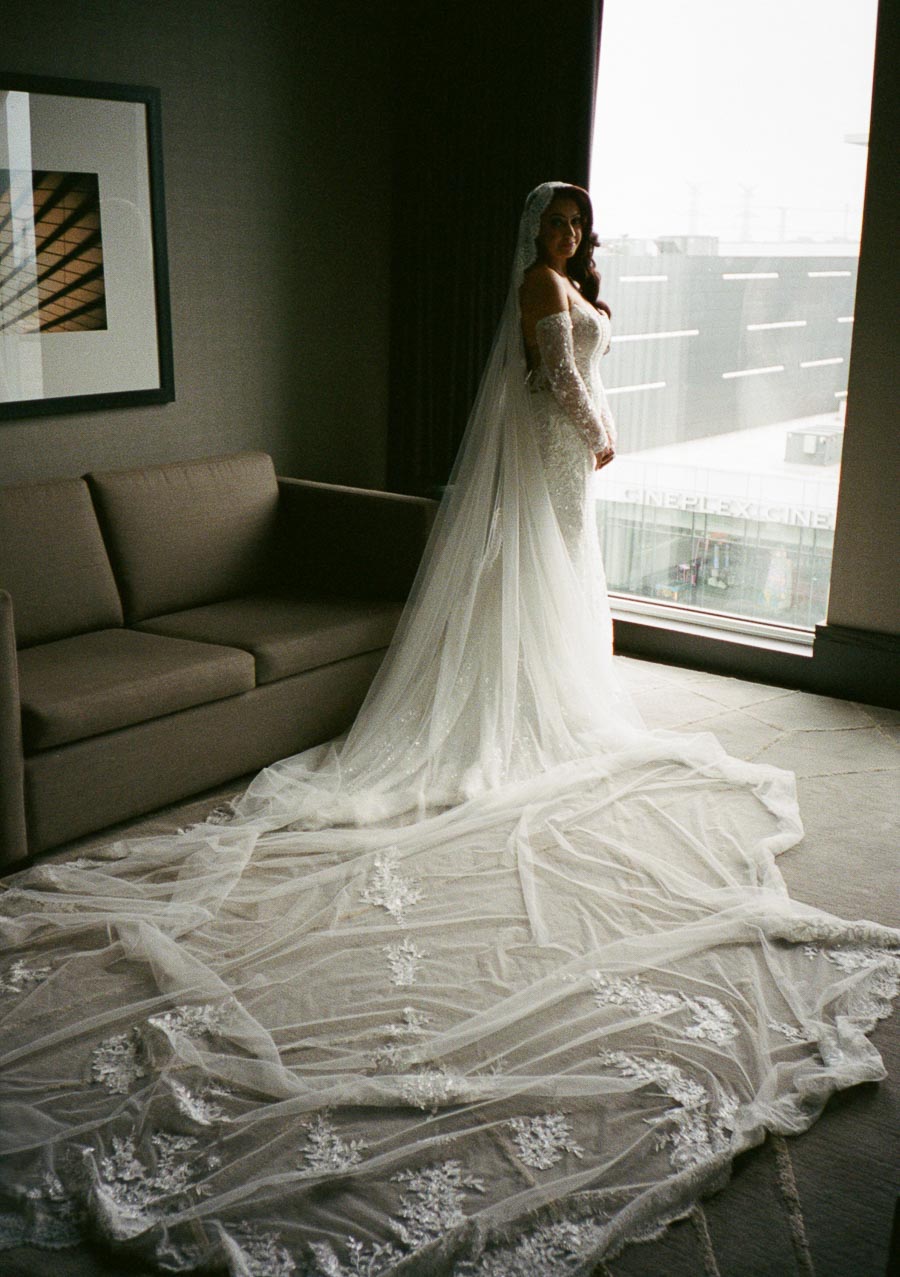Bride in elegant long-sleeve lace wedding gown with flowing train standing by a window in a modern room.