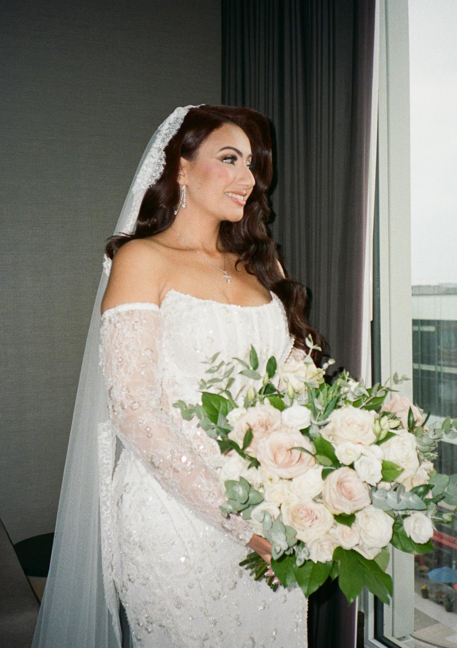 Bride in elegant white gown holding a bouquet of pink and white roses, standing by a window with a soft smile.