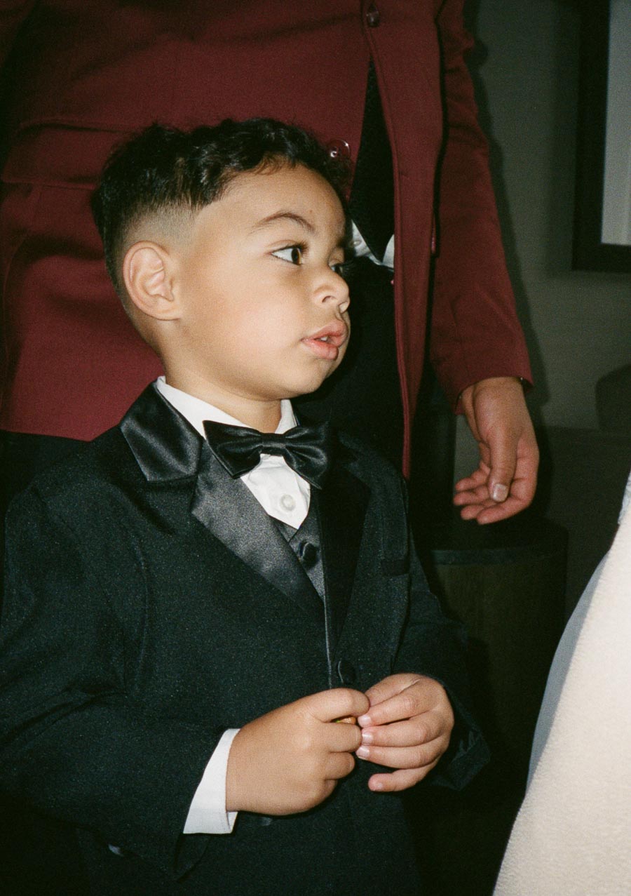 A young child dressed in a formal black tuxedo with a bow tie at an event, standing next to an adult in a burgundy outfit.