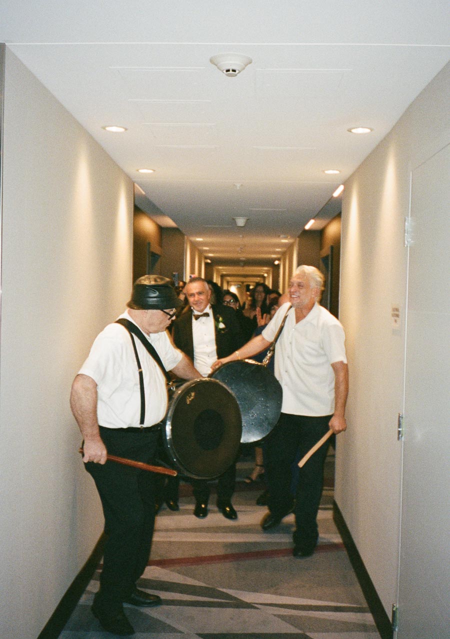 A joyful celebration in a hallway, featuring two elderly men playing large drums, with one man in a tuxedo smiling and a crowd in the background.