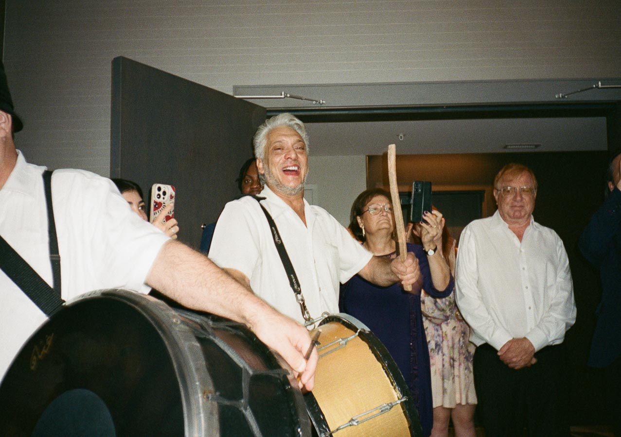 A lively celebration featuring a joyful drummer leading a group of people in a festive setting, with attendees capturing the moment on smartphones.