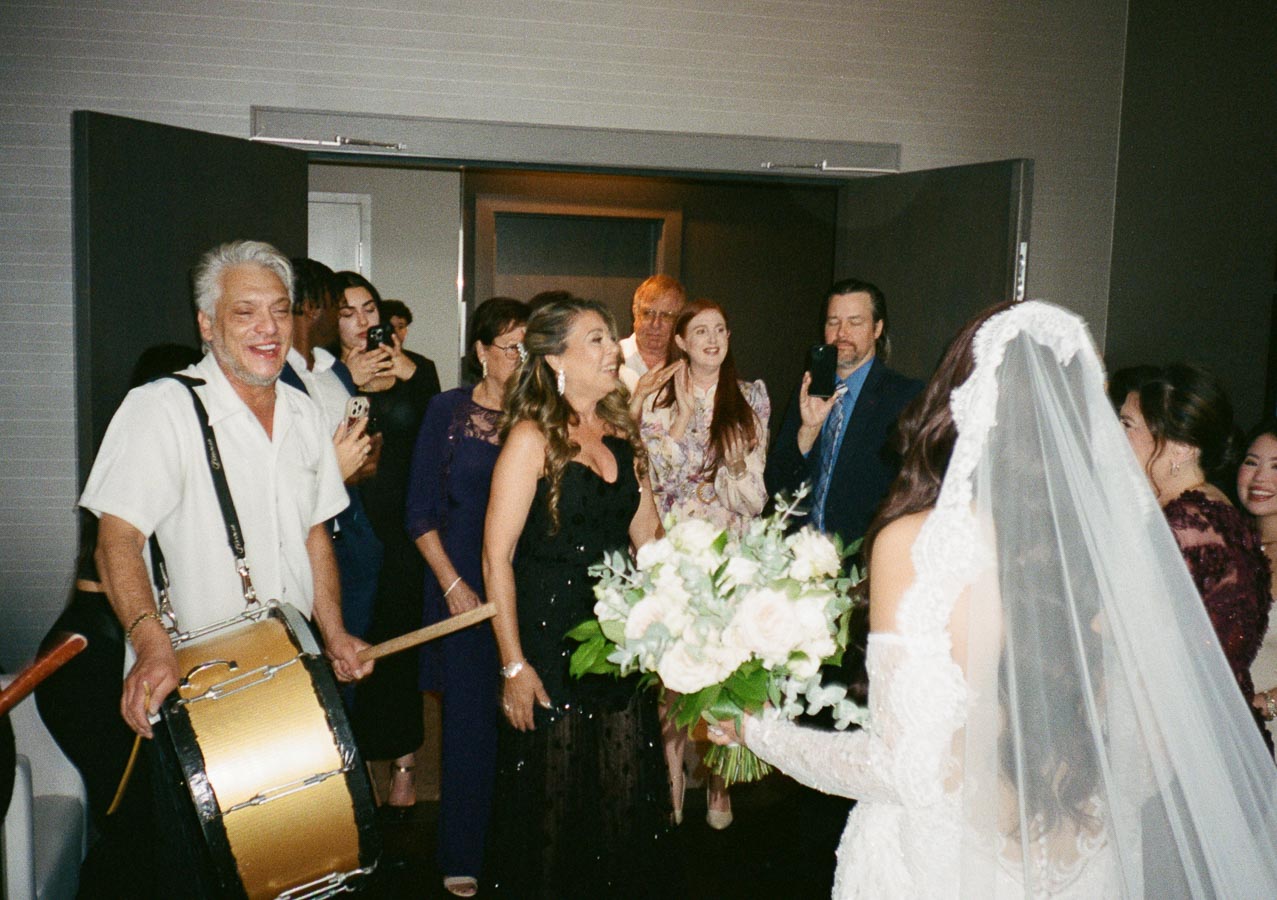 A joyful wedding celebration with a smiling bride in a white lace dress and veil holding a bouquet, surrounded by cheering guests. A man with a drum leads the group, capturing a moment of festivity and happiness.