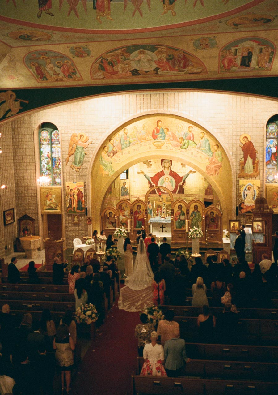 A bride and groom stand at the altar during a traditional church wedding ceremony, with ornate religious artwork and stained glass windows in the background, surrounded by seated guests.
