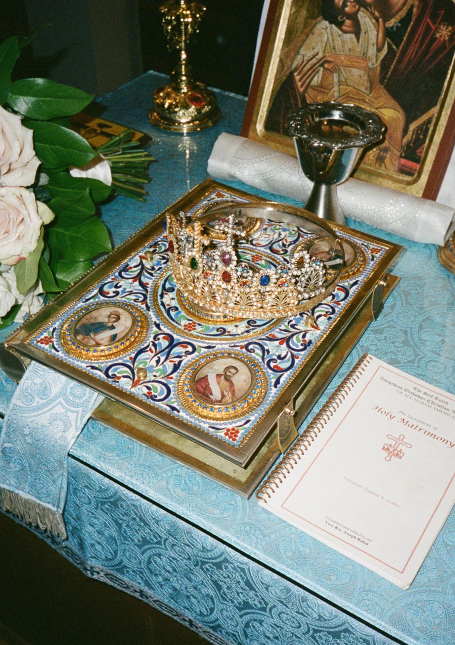 Intricately decorated religious altar with a jeweled crown, ornate book with iconography, white roses, and a Holy Matrimony booklet on a blue patterned cloth.