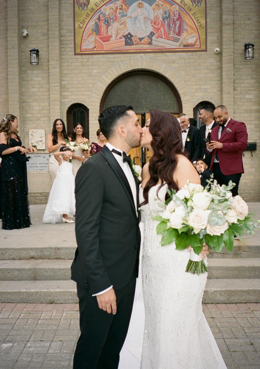 Bride and groom share a kiss outside a church with wedding party celebrating in the background.