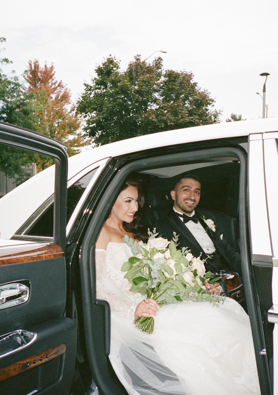 A bride and groom smiling inside a luxurious car, with the bride holding a bouquet of white and green flowers, dressed in an elegant wedding gown, and the groom in a black tuxedo.