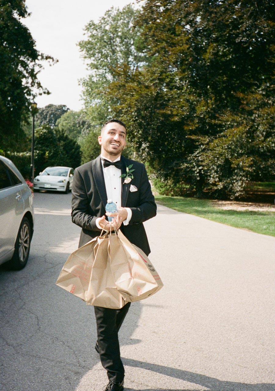 A man in a black tuxedo carrying paper shopping bags and a water bottle, walking along a tree-lined pathway near parked cars.