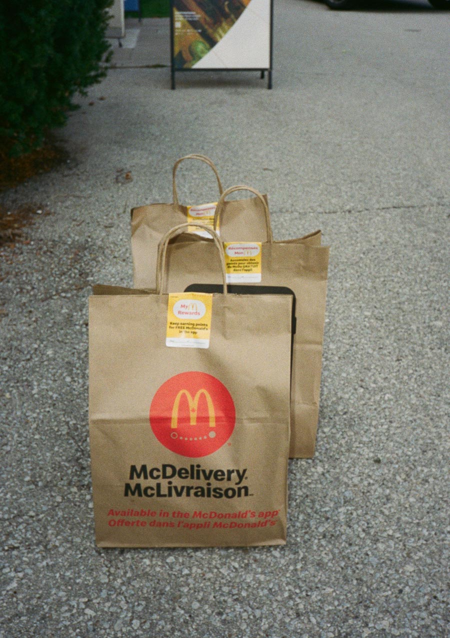McDonald's brown paper delivery bags on a sidewalk, featuring the McDelivery logo and promotional labels, emphasizing food delivery service.