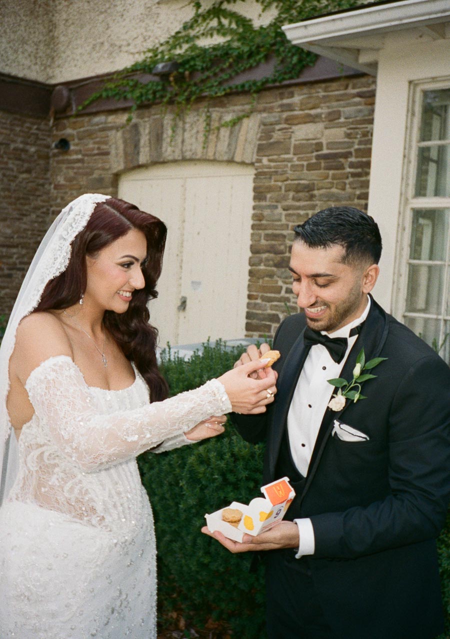 A bride and groom in wedding attire share a playful moment eating chicken nuggets outside a rustic building.