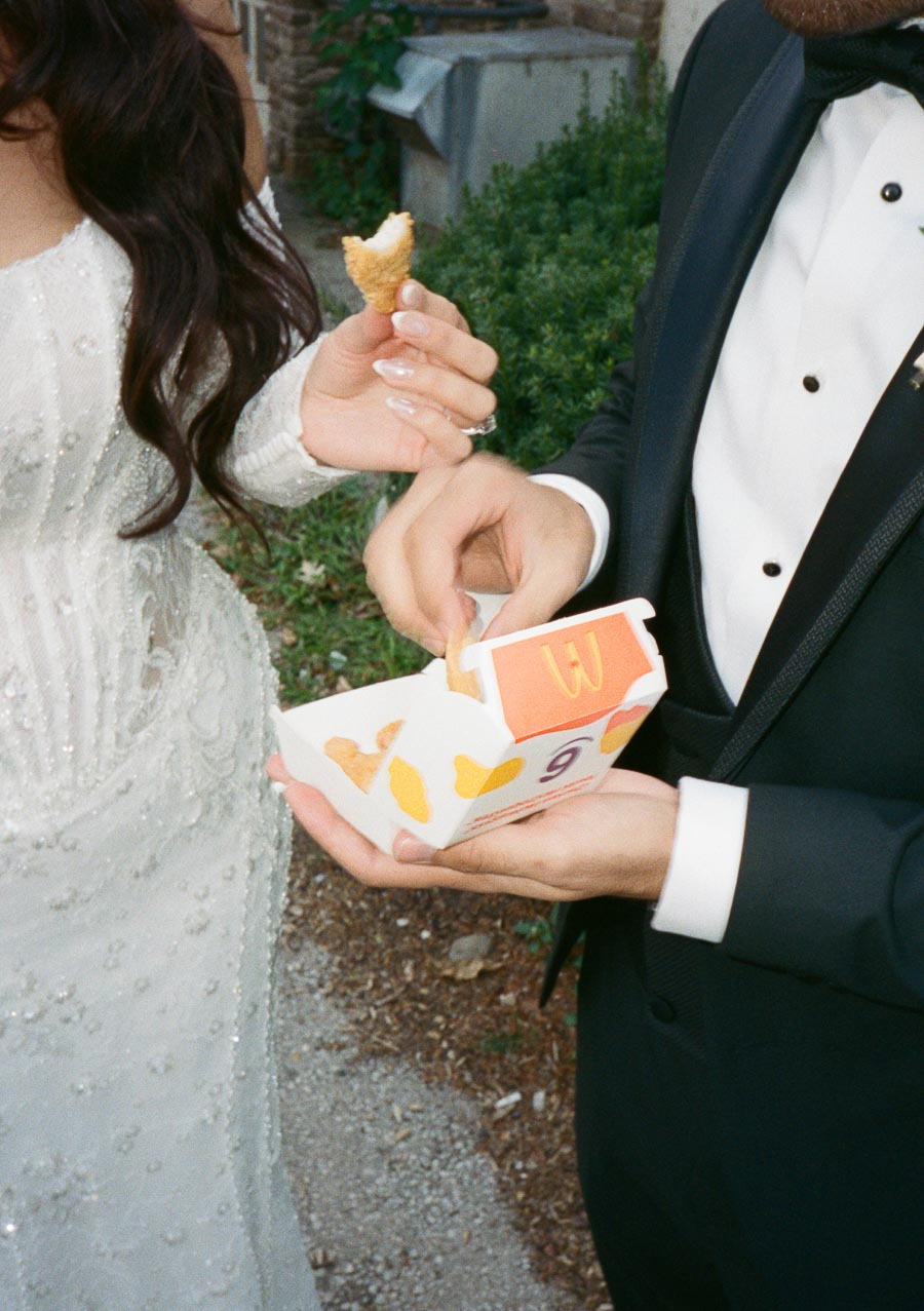 A bride in a white wedding dress and a groom in a black tuxedo enjoy fast food chicken nuggets together outdoors.