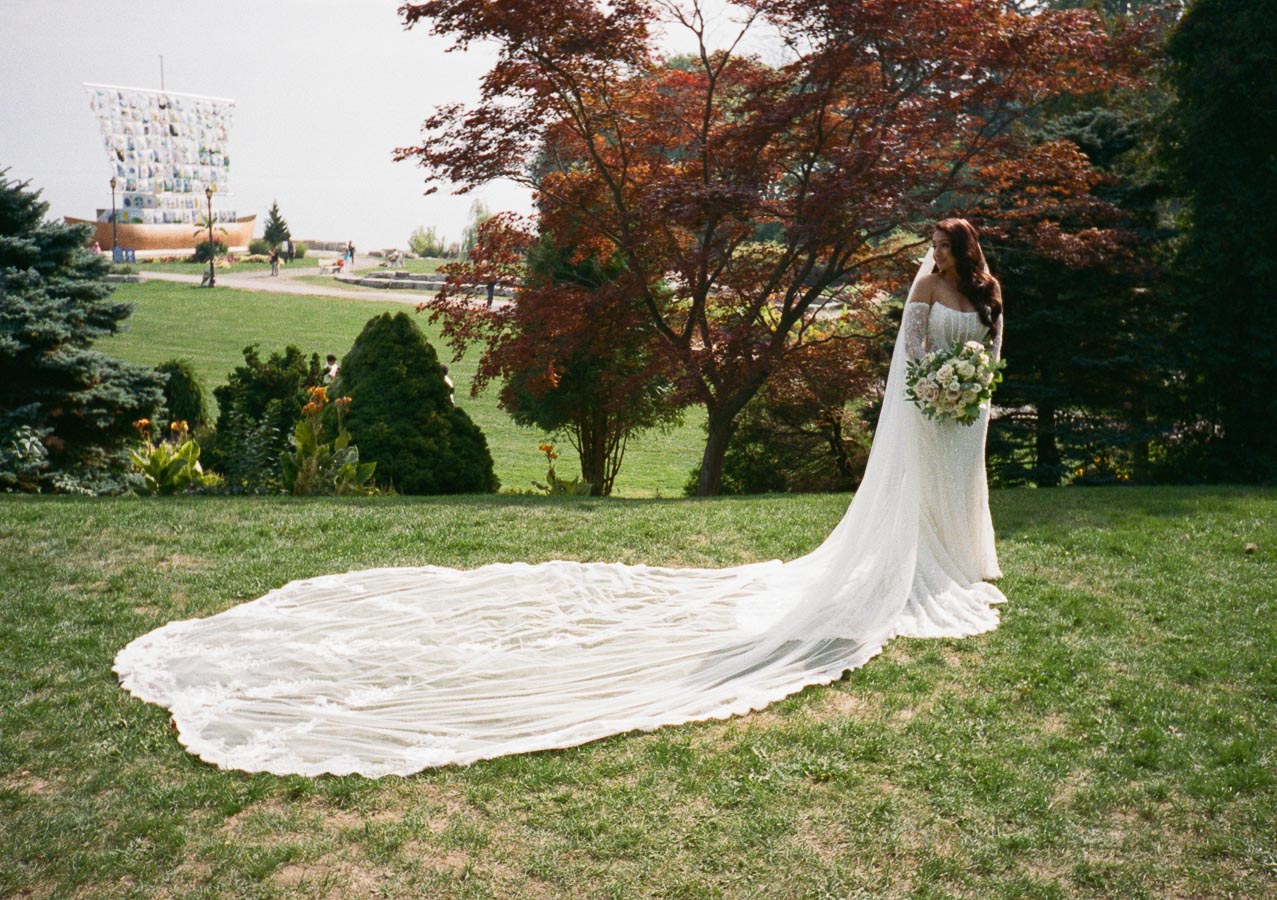 A bride in a flowing white gown and long train stands gracefully on a lush green lawn, holding a bouquet of white and blush flowers, with vibrant autumn trees highlighting the serene outdoor setting.