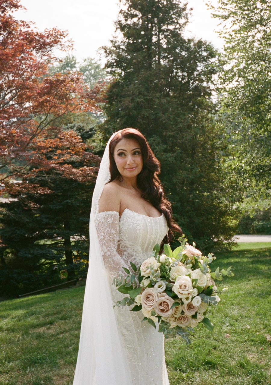 Beautiful bride in a lace wedding gown holding a bouquet of roses, standing outdoors in a lush garden setting.