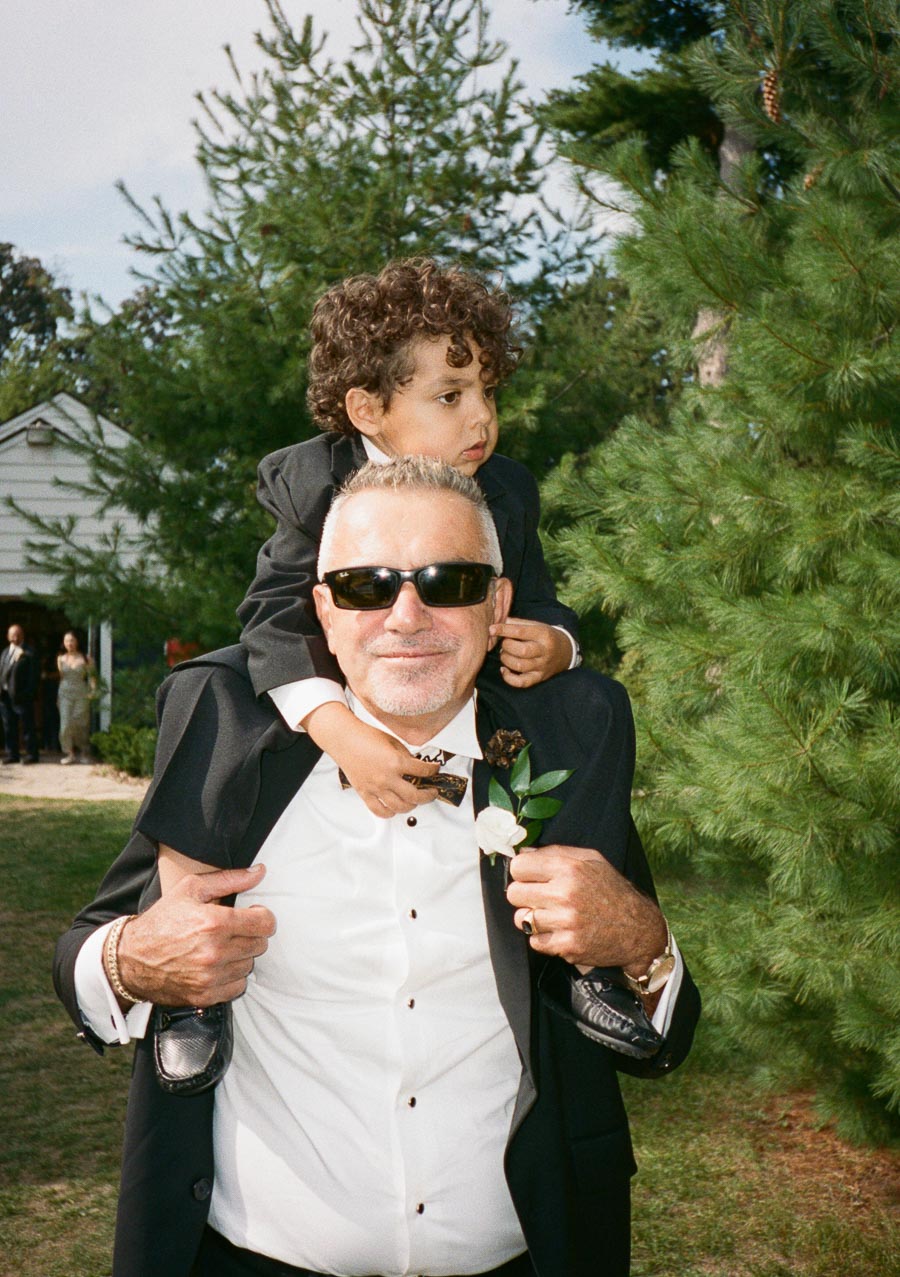 Elderly man wearing sunglasses and a tuxedo carries a young boy on his shoulders in a garden setting, surrounded by trees and a building in the background.