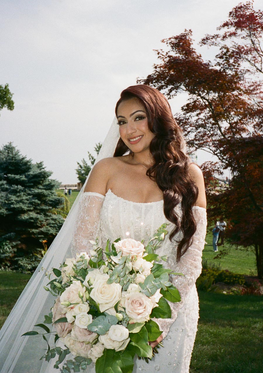 A bride in an elegant white wedding gown holds a lush bouquet of white and pink roses, standing outdoors with a backdrop of green trees and grass.