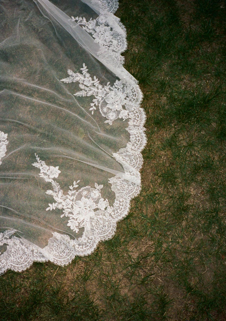 Close-up of an intricate white lace veil with floral patterns draped over green grass, highlighting delicate embroidery and wedding elegance.