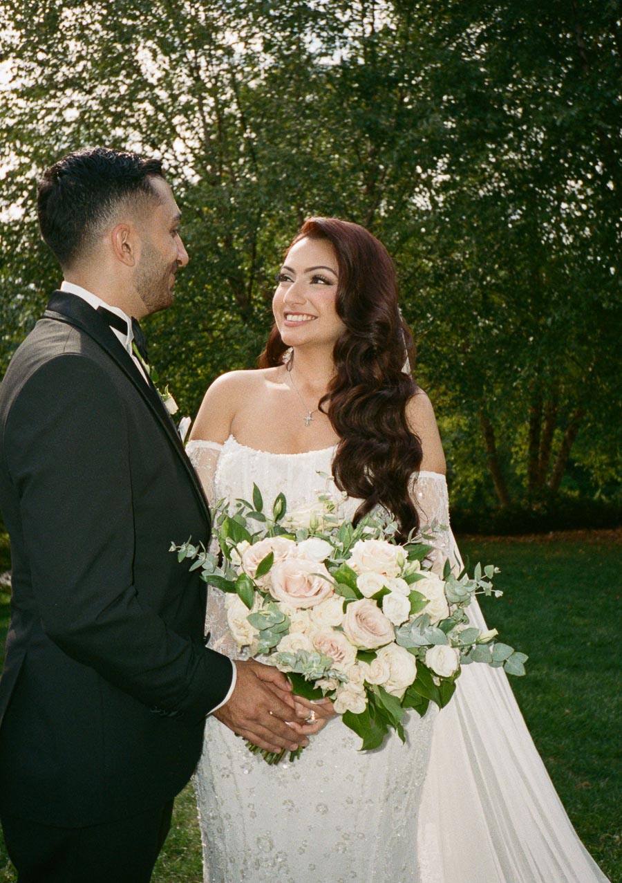 A happy couple in wedding attire gazing at each other, with the bride holding a bouquet of pink and white roses, set against a lush green background.