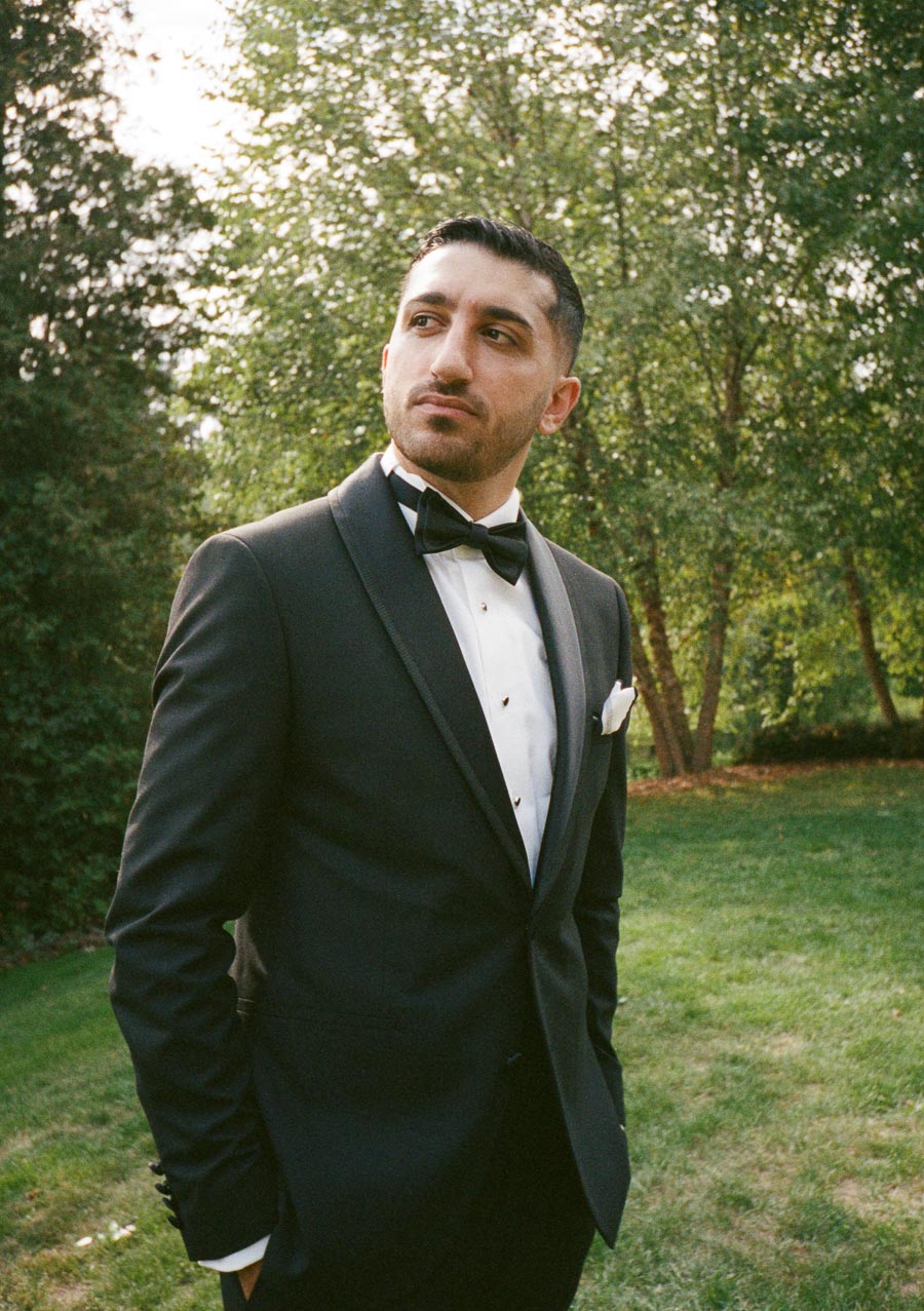 A man in a black tuxedo with a bow tie stands in a garden setting, surrounded by greenery, appearing thoughtful and confident.