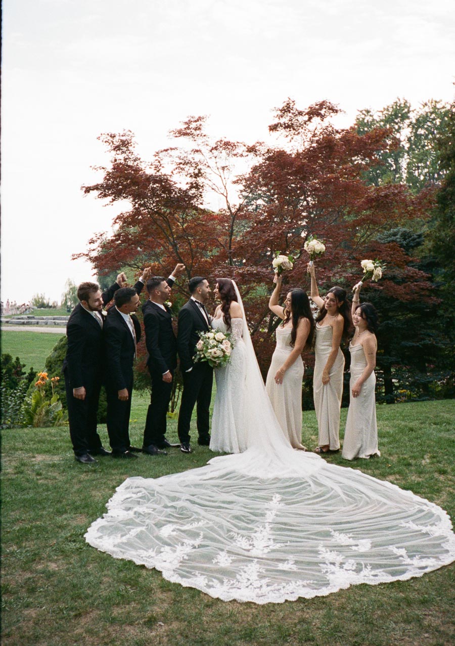 A newlywed couple celebrates with their wedding party outdoors. The bride, in a long white gown with an extended veil, holds a bouquet and stands beside the groom, who is in a black suit. Bridesmaids in light dresses and groomsmen in dark suits celebrate with raised bouquets against a backdrop of trees and greenery.