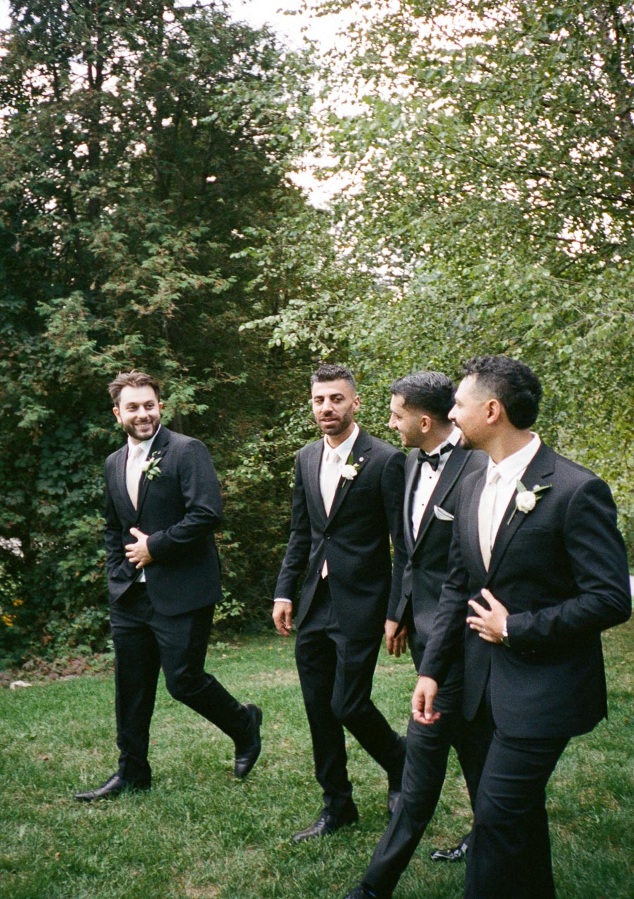 Four groomsmen in sleek black suits walk outdoors on a lush green lawn, surrounded by trees, enjoying a light-hearted moment before a wedding ceremony.