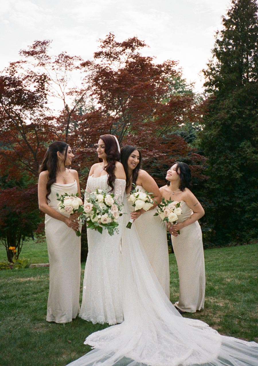 A bride in a white wedding gown stands with three bridesmaids in beige dresses, holding bouquets of white and pink roses, set against a lush garden backdrop with red and green foliage.