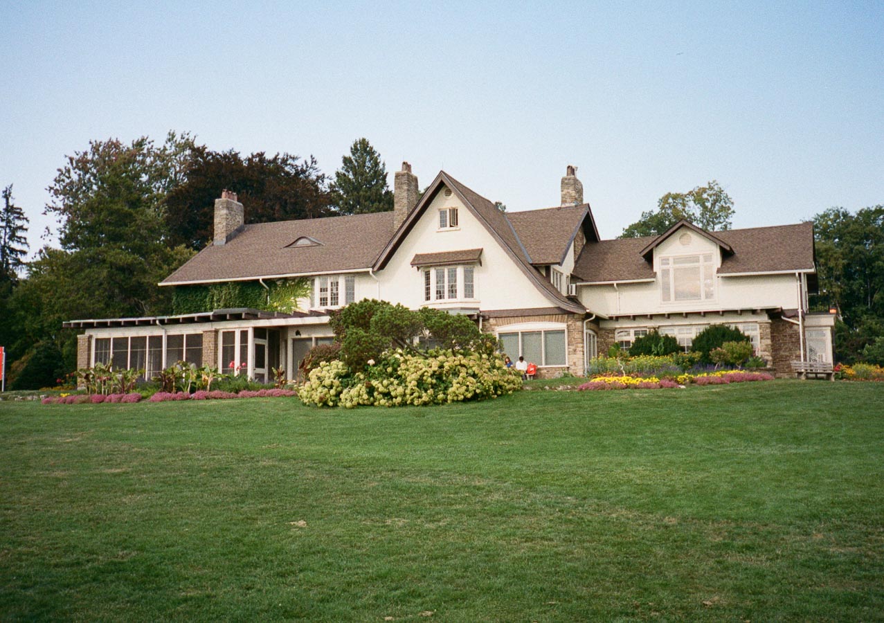 Large traditional countryside house with a brown roof and stone chimneys, surrounded by lush green lawn and colorful flower beds in a serene outdoor setting.