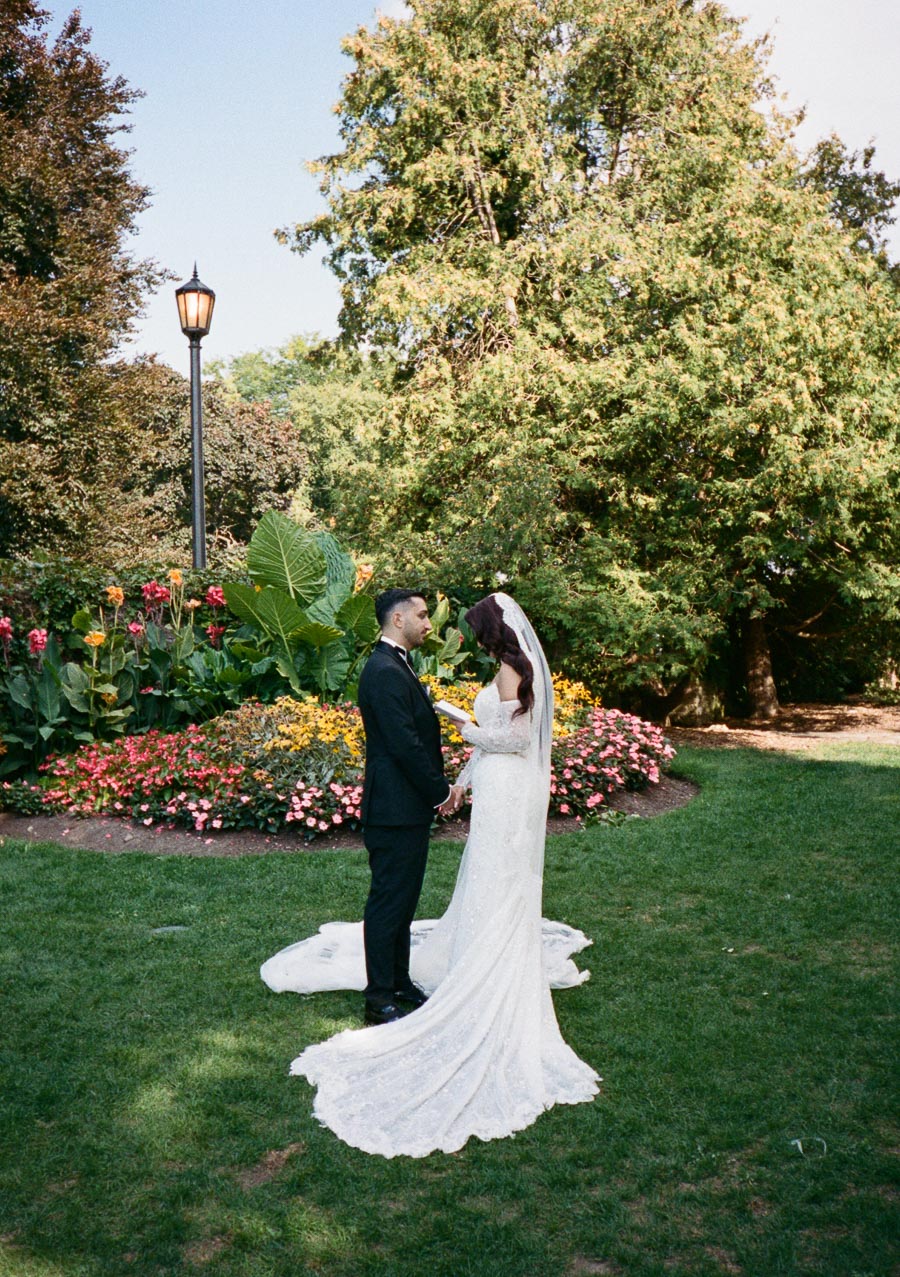 A bride and groom exchanging vows in a lush garden setting, surrounded by vibrant flowers and greenery, under the early afternoon sky.