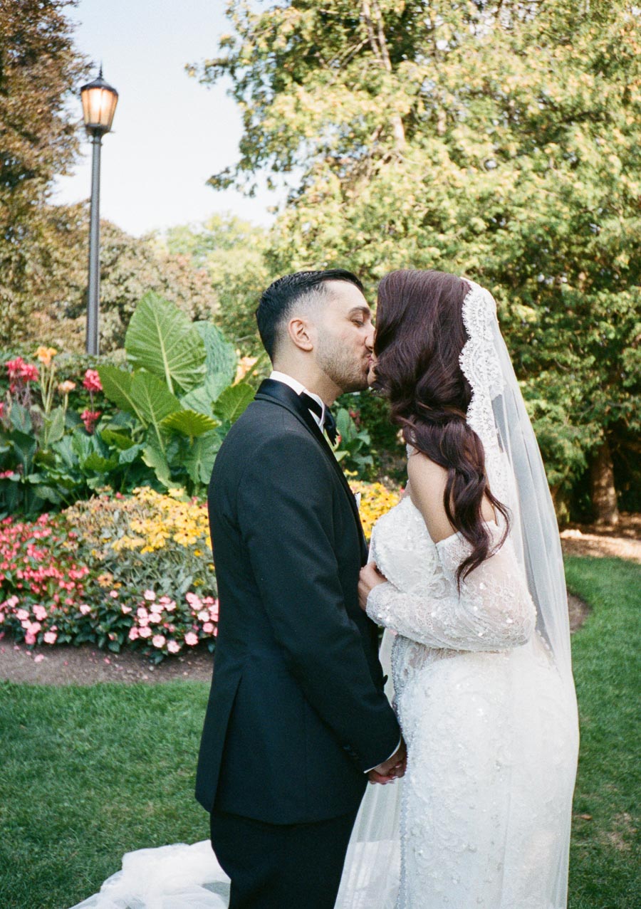 A bride and groom share a romantic kiss in a lush garden setting, surrounded by vibrant flowers and greenery, on their wedding day.