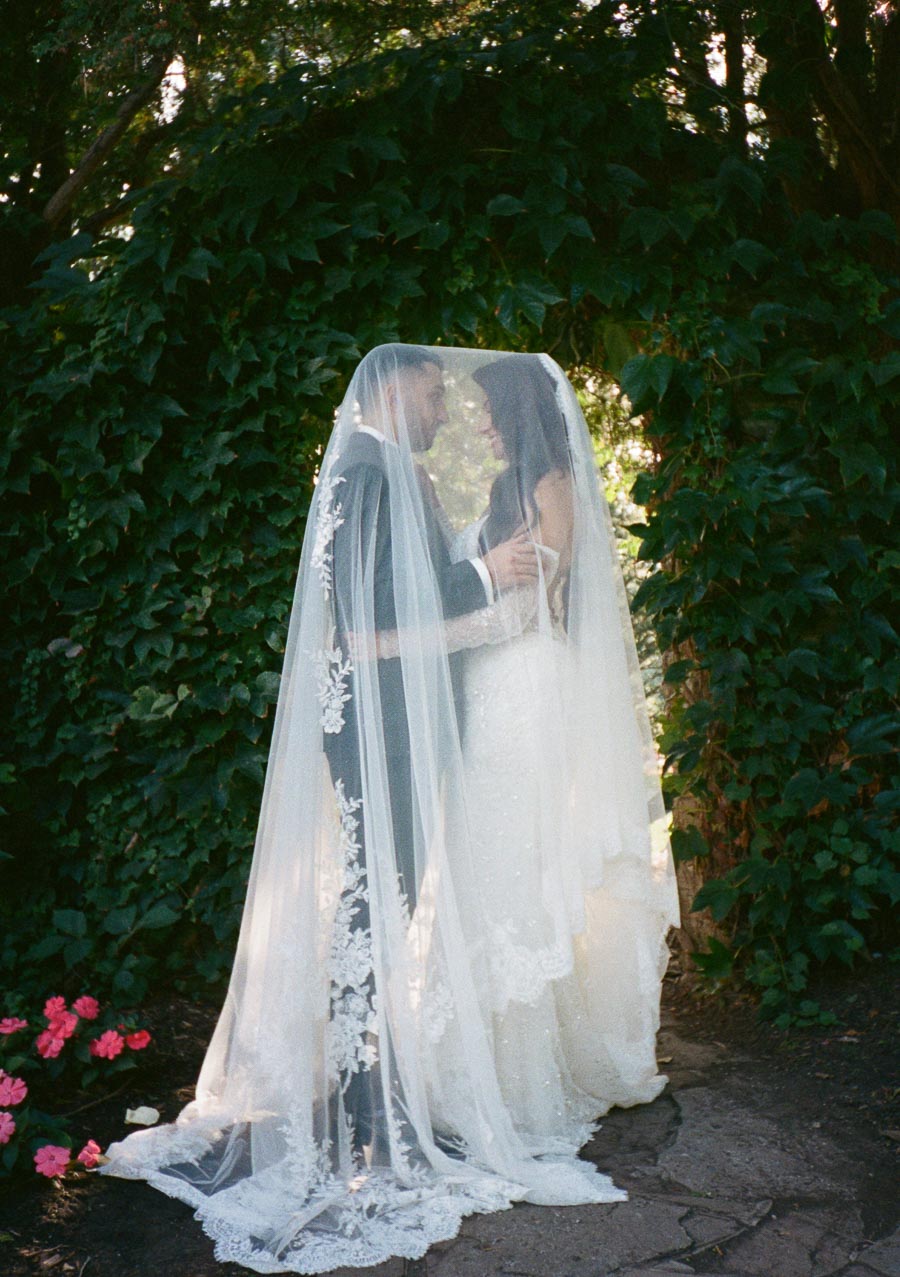 Wedding couple embracing under a sheer veil in a lush garden setting with green foliage and pink flowers, capturing a romantic and intimate moment.