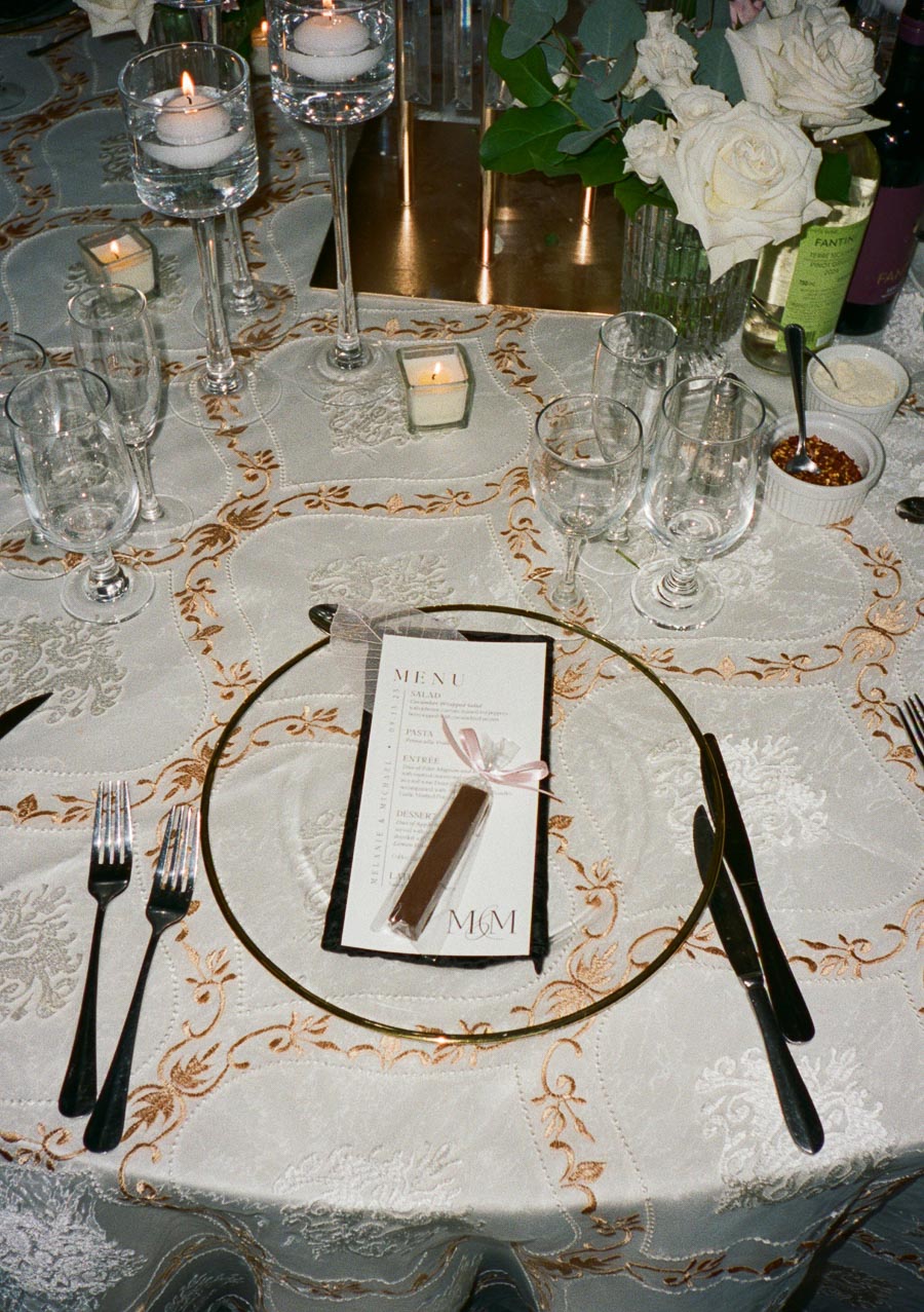 Elegant table setting at a formal event, featuring a decorative menu placed on a gold charger, surrounded by wine glasses, cutlery, floating candles, and a vase of white roses on an ornate tablecloth.