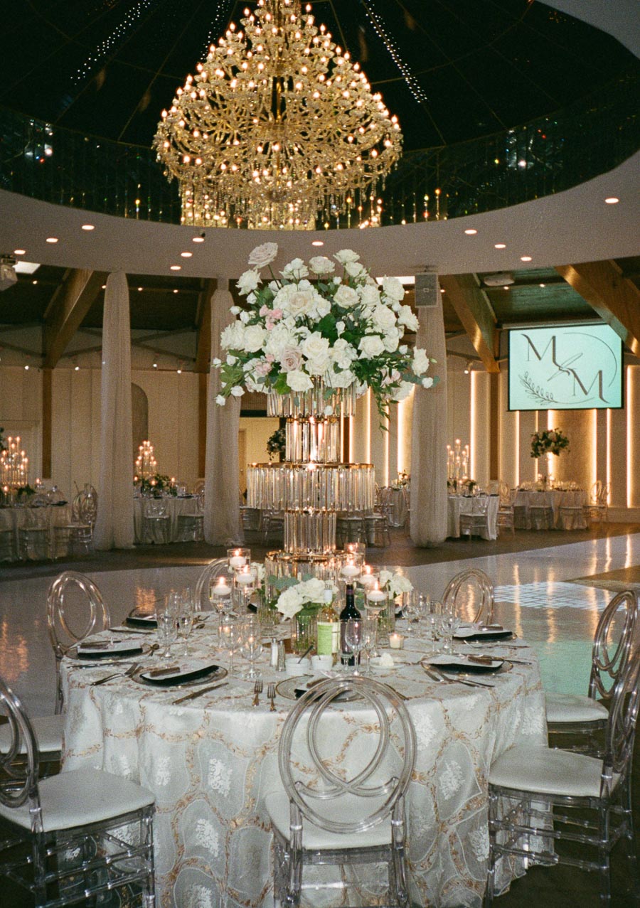 Elegant wedding reception with a beautifully set round table adorned with white floral centerpieces and lit candles, under a grand crystal chandelier in a luxurious venue.