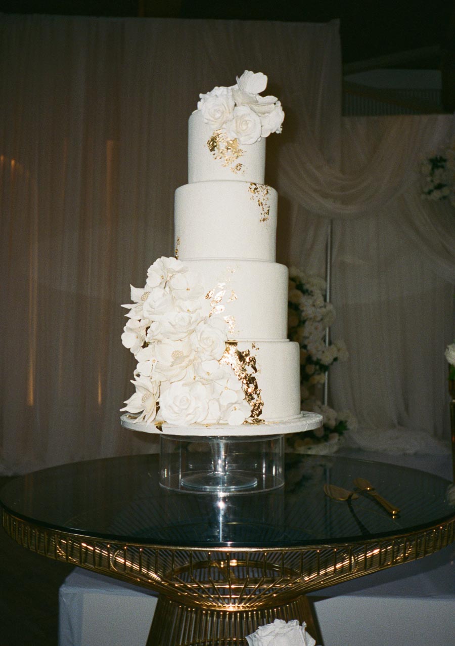 Four-tier white wedding cake adorned with elegant white floral decorations and gold leaf accents, displayed on a stylish glass and gold table against a draped fabric backdrop.