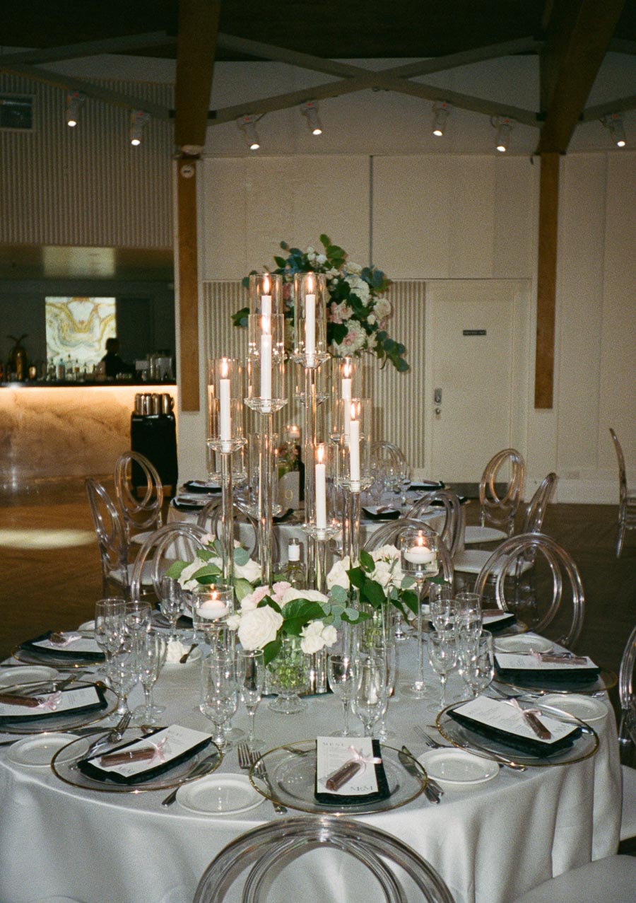 Elegant wedding reception table setting with tall candle centerpiece, floral arrangements, and clear chairs in a sophisticated venue.