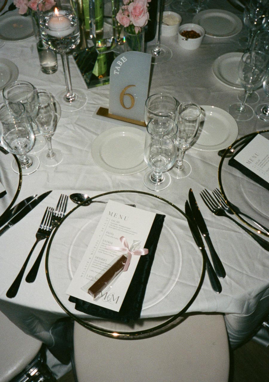 Elegant wedding table setting with white tablecloth, glassware, and floral centerpiece. Menu and chocolate favor tied with pink ribbon on plate, marked Table 6.