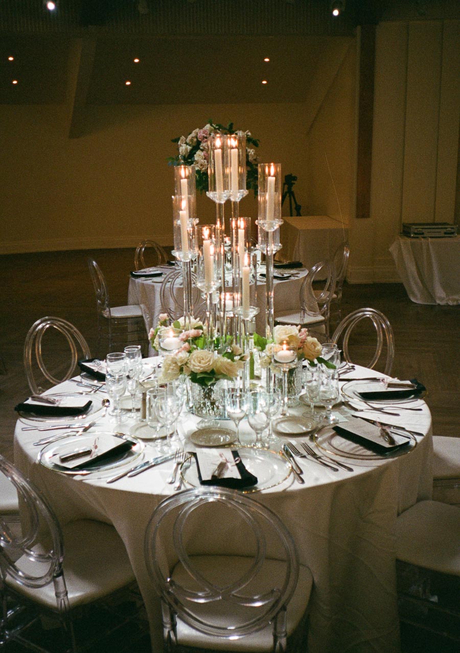 Elegant round table setup with tall candle centerpiece, surrounded by clear, modern chairs. The table is adorned with white linens, floral arrangements, and neatly arranged cutlery and glassware, creating a sophisticated ambiance suitable for weddings or formal events.