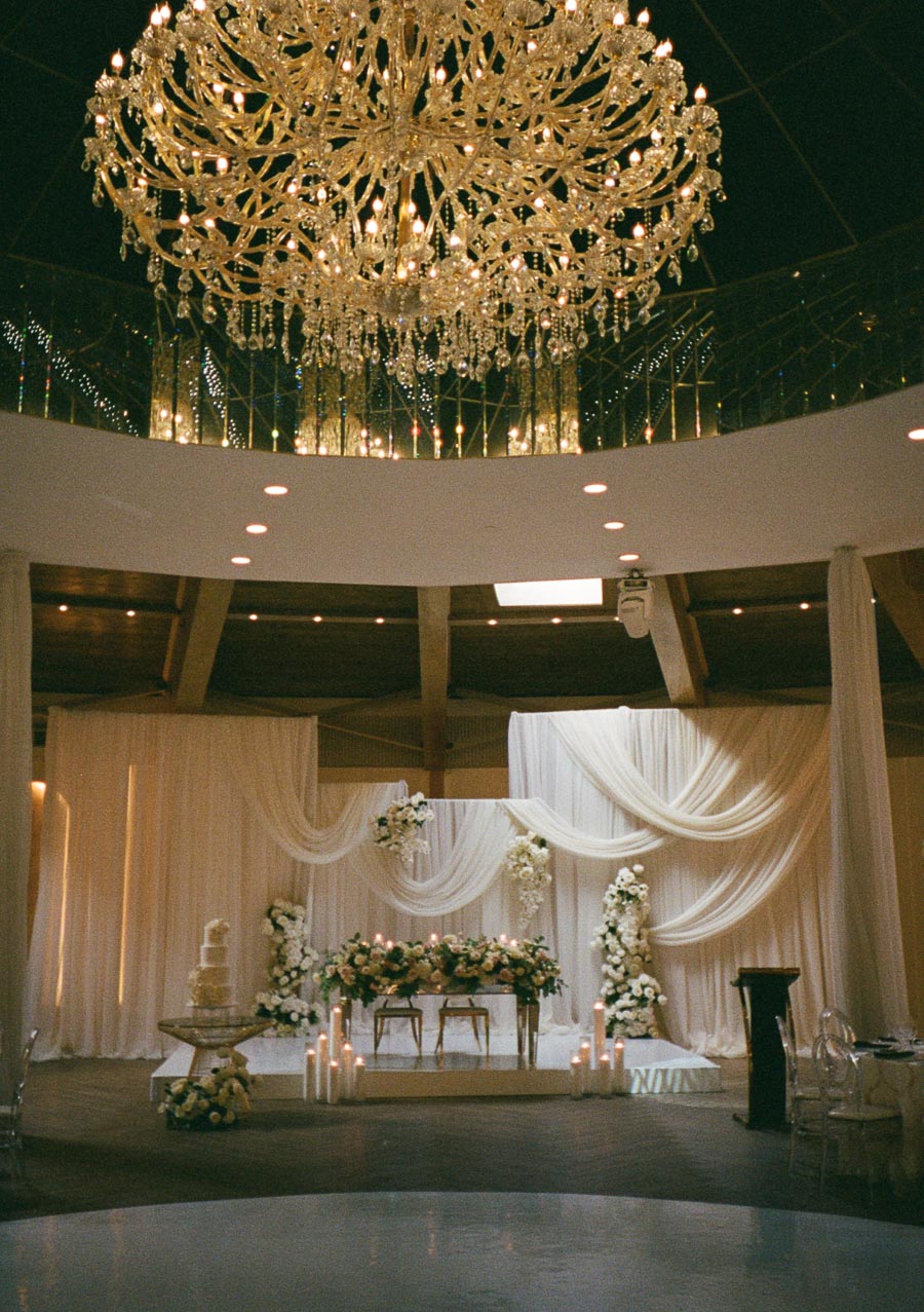 Elegant wedding reception venue with a grand chandelier, draped fabric backdrop, floral arrangements, and a tiered cake, creating a luxurious and romantic atmosphere.