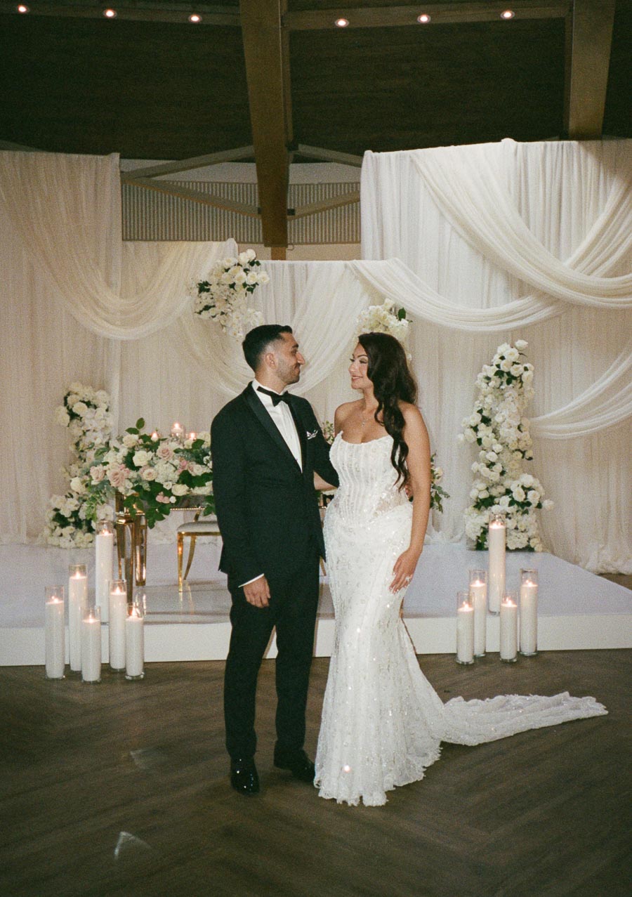 Elegant wedding couple gazing at each other with love, surrounded by a beautifully decorated venue with white drapery, floral arrangements, and candlelight.