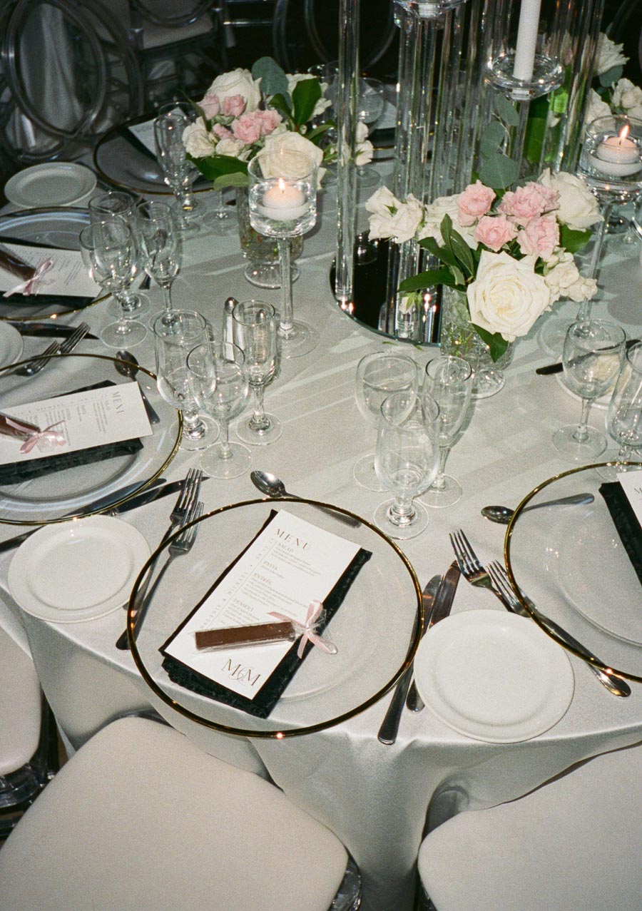 Elegant wedding table setting with glass plates, silverware, crystal glasses, white floral centerpiece with pink accents, candles, and personalized menus on a white tablecloth.