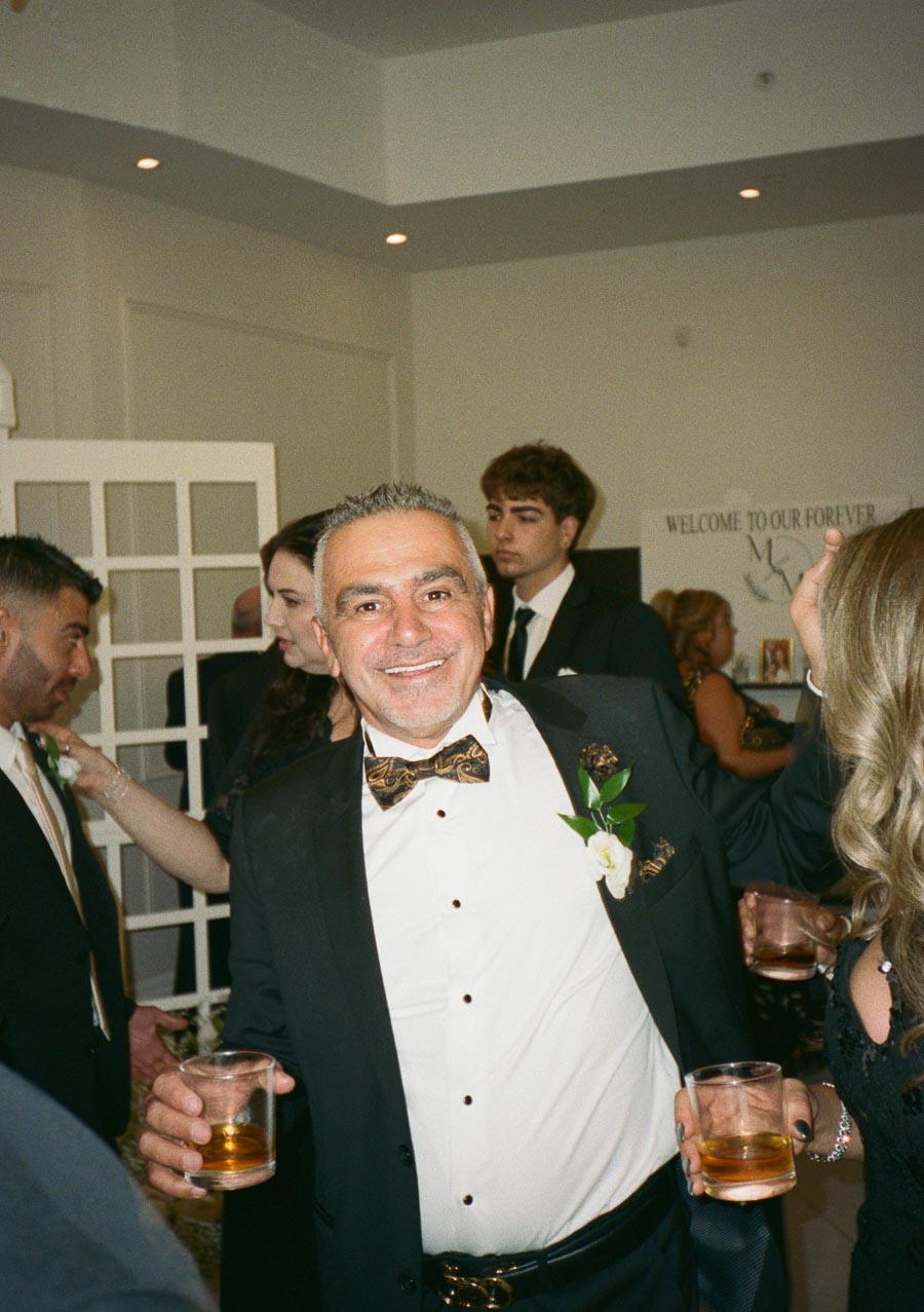 Man in a suit smiling at a wedding reception, holding a drink, with a boutonniere and a patterned bow tie, surrounded by other guests in formal attire.