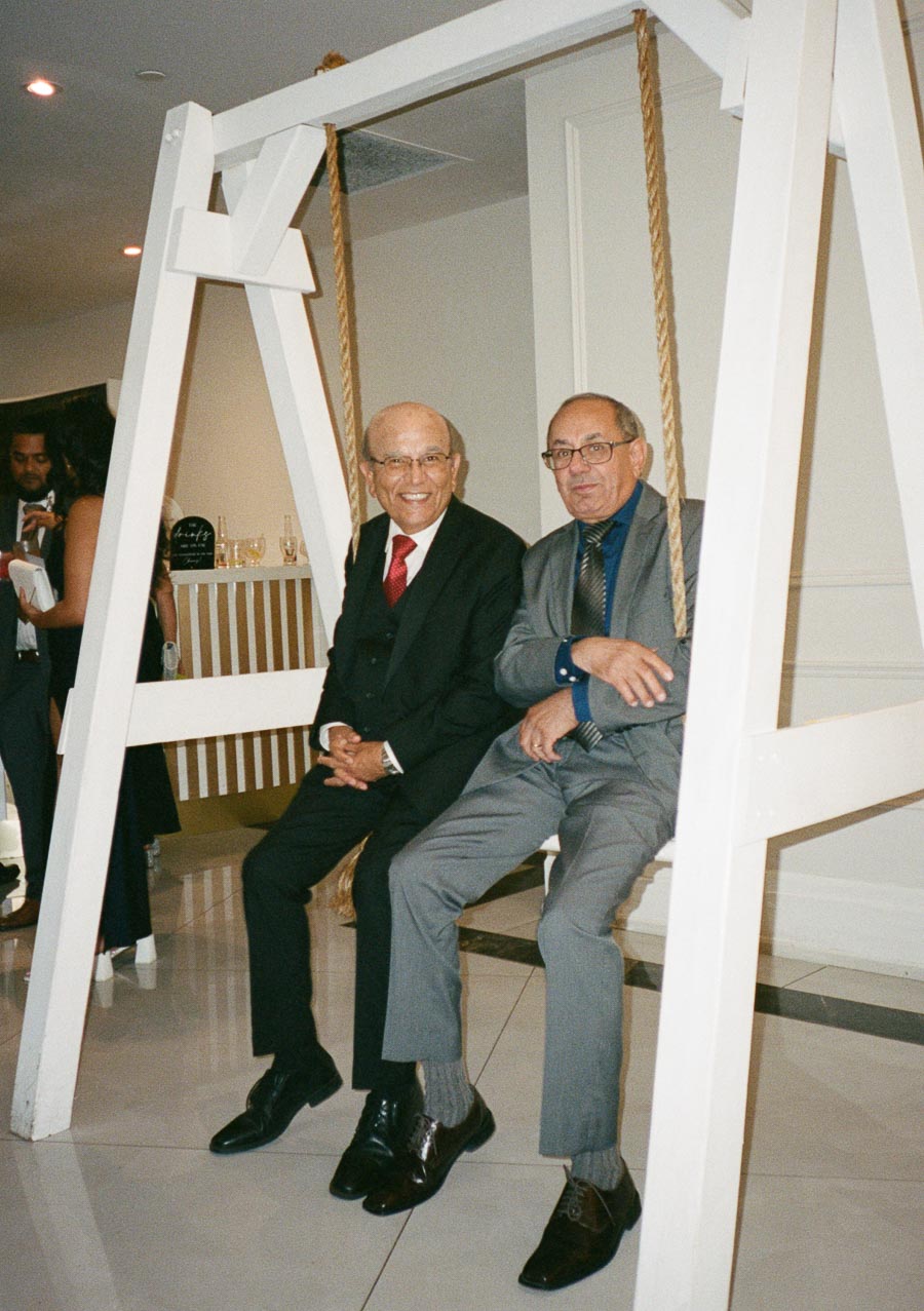 Two elderly men in suits sitting on a swing indoors, smiling at a social event with people in the background.