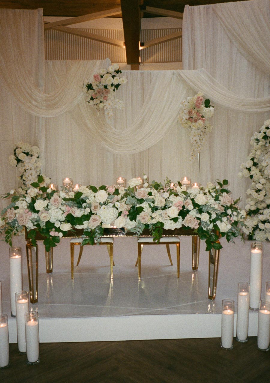 Elegant wedding reception table adorned with lush white and blush floral arrangements, surrounded by glowing candles, set against a backdrop of draped white fabric and romantic flower accents.