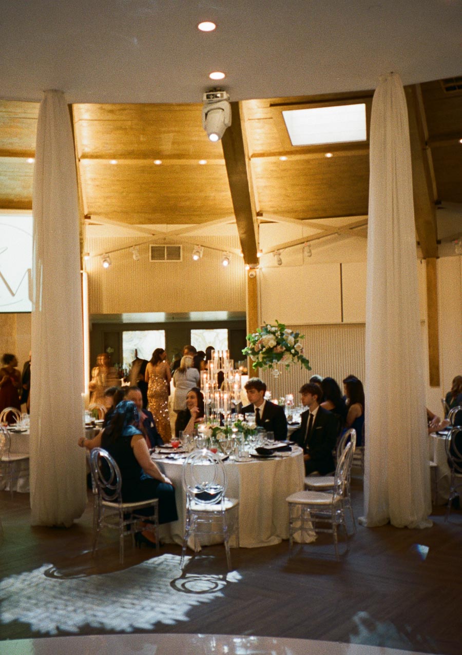 Elegant wedding reception in a beautifully decorated hall with guests seated around a table adorned with floral arrangements and candle centerpieces.