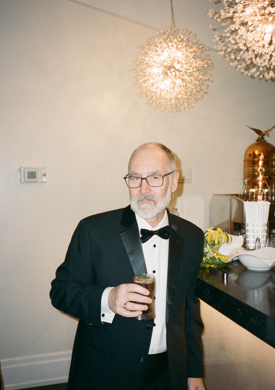 A man in a formal tuxedo holding a drink, standing by a bar adorned with elegant decor and a bright chandelier overhead.
