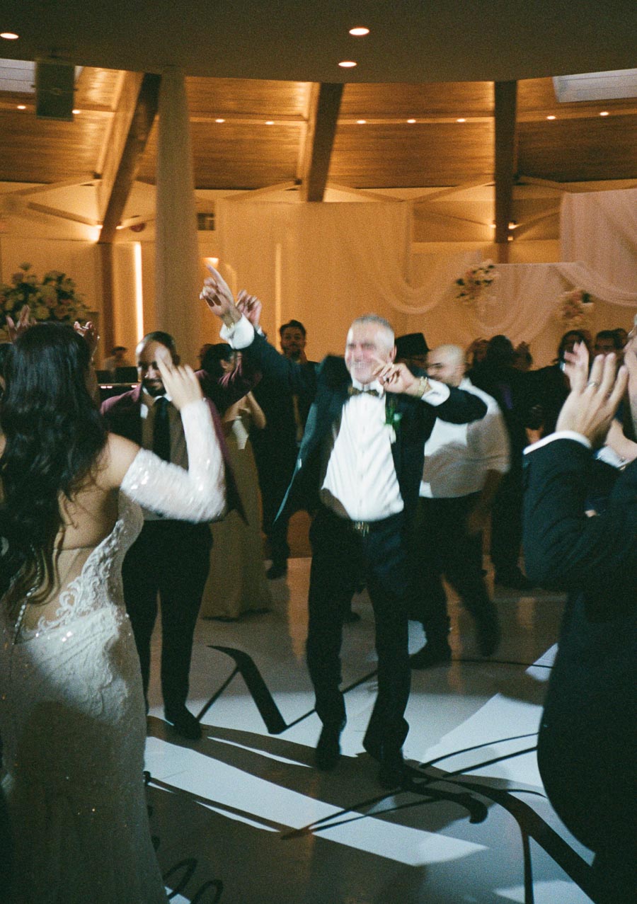A lively wedding reception with guests dancing energetically on the dance floor, featuring a bride in an elegant lace gown and a groom in a tuxedo amidst a warmly lit venue.