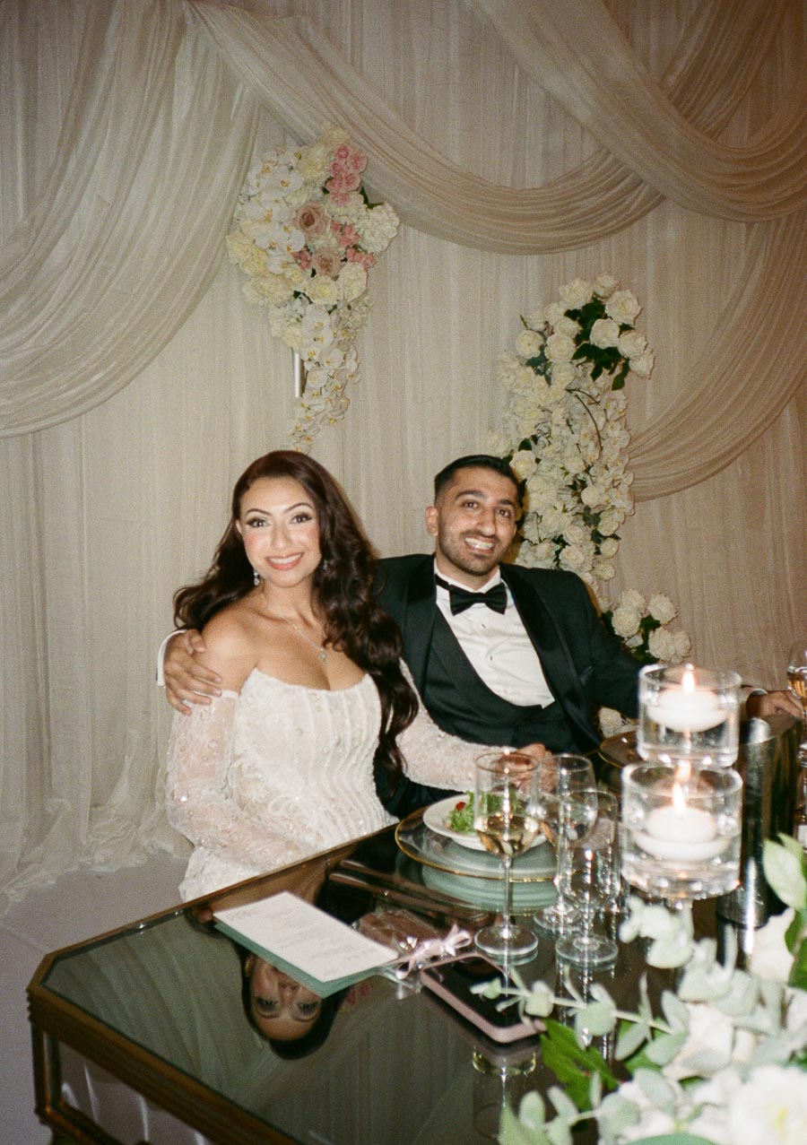 Bride and groom smiling while seated at a decorated wedding reception table with floral arrangements and candles.