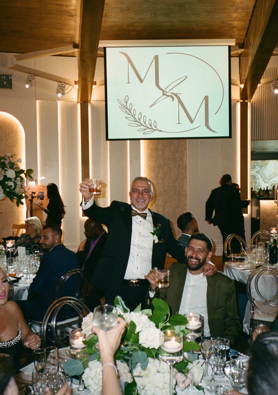 A joyful wedding reception scene with elegantly dressed guests toasting at a beautifully decorated table adorned with white floral centerpieces and candles, beneath a projected monogram display.