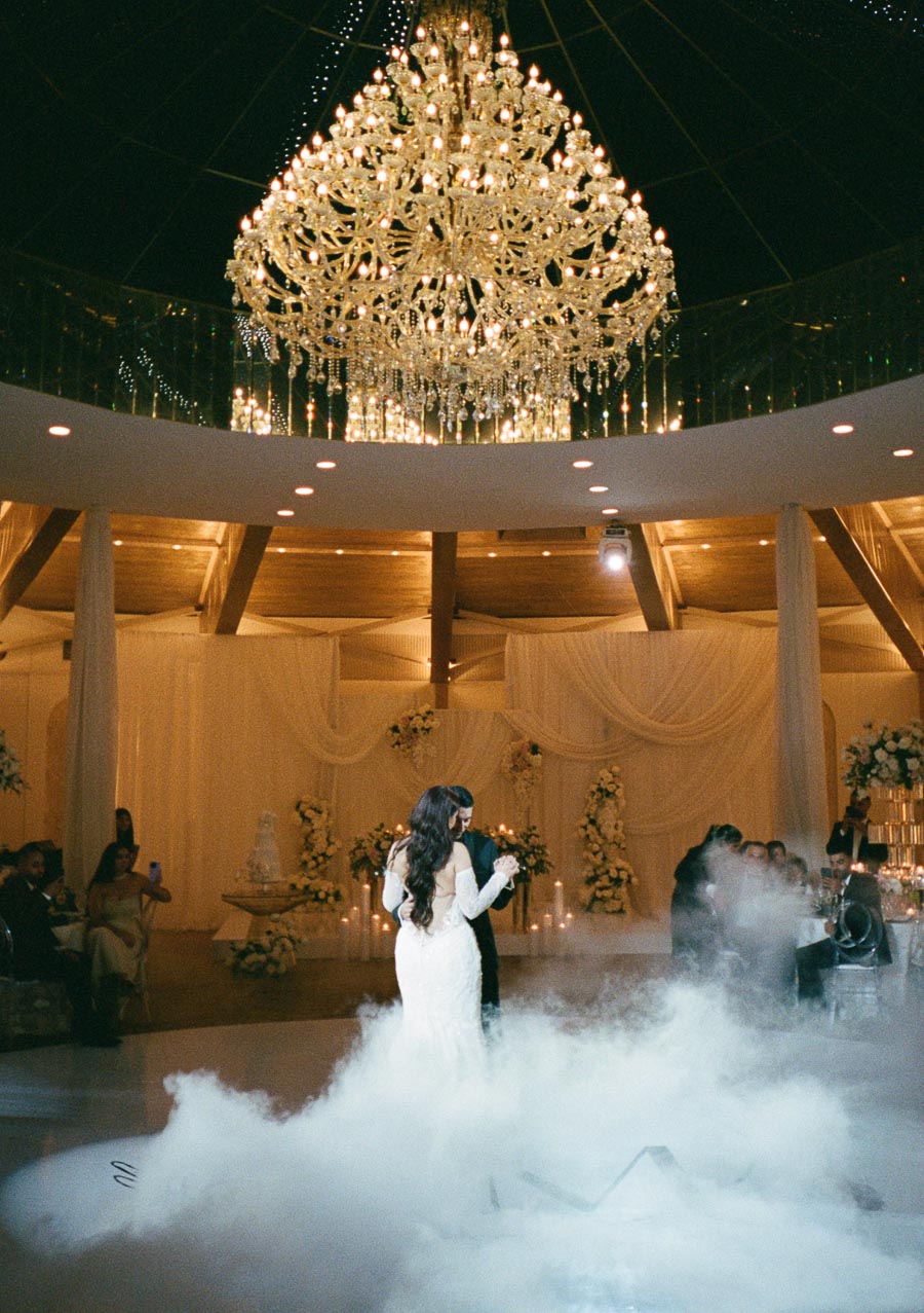 Elegant wedding couple sharing their first dance under a magnificent crystal chandelier, surrounded by a romantic atmosphere with soft lighting and decorative flowers.