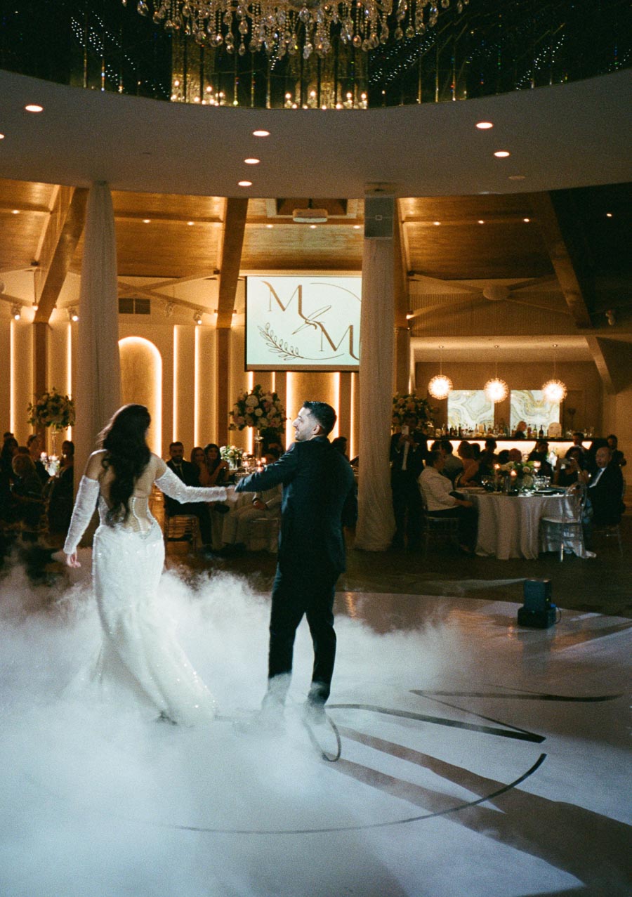 A bride and groom dance on a mist-covered floor in an elegantly decorated wedding venue, with ambient lighting and a monogrammed backdrop.
